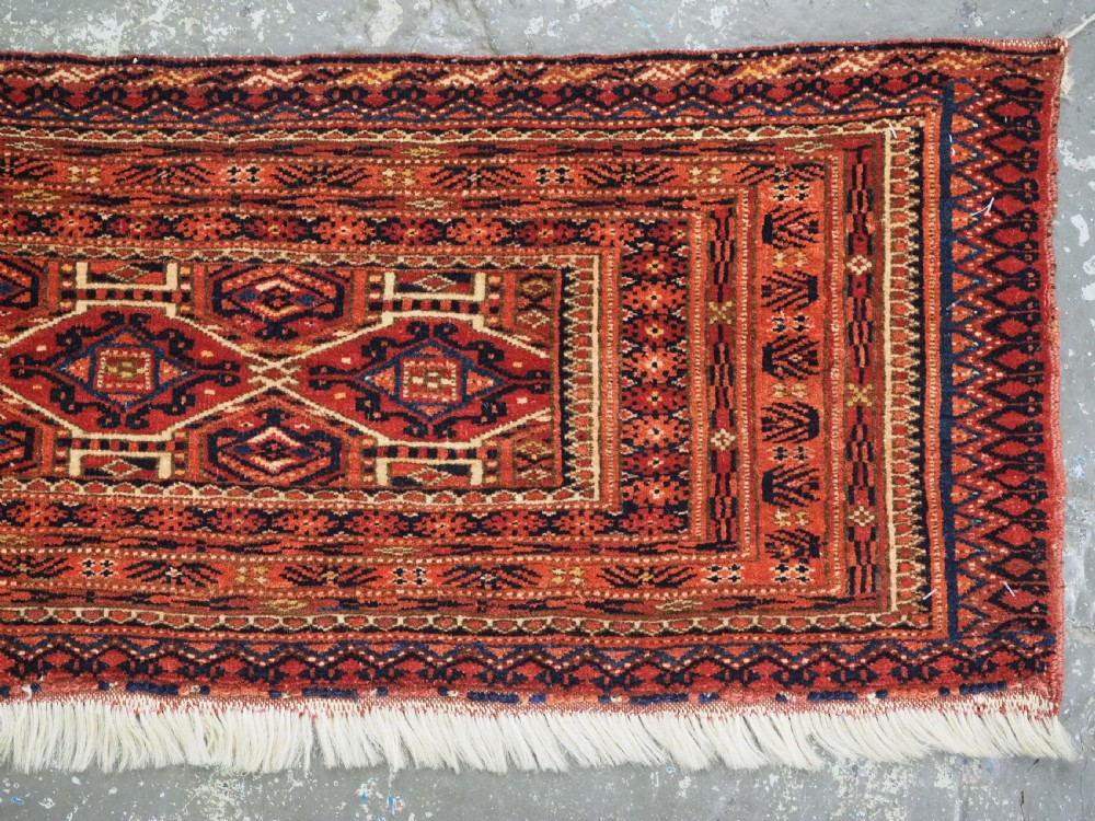An elongated antique Turkmen torba textile displaying geometric diamond medallion motifs in deep red, dark navy, and cream wool with fringed bottom edge. View V