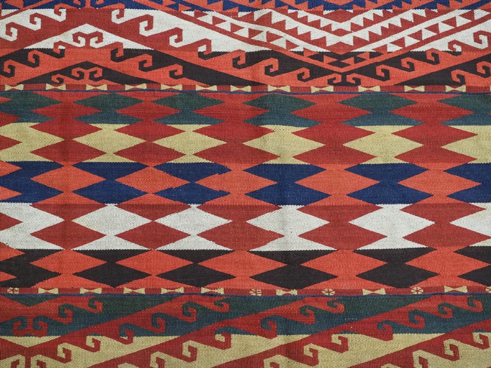 A rectangular handwoven flatweave kilim panel featuring horizontal bands of geometric diamonds, zig-zags, and latch-hook motifs in red, cream, navy blue, and ochre wool. View IV