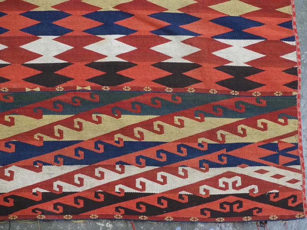 A rectangular handwoven flatweave kilim panel featuring horizontal bands of geometric diamonds, zig-zags, and latch-hook motifs in red, cream, navy blue, and ochre wool. View V
