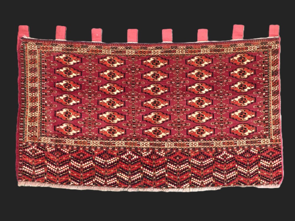 A woven deep red antique Turkmen chuval face textile featuring 30 repeating octagonal guls, an ornate geometric lower border, and fabric hanging loops along the top edge.