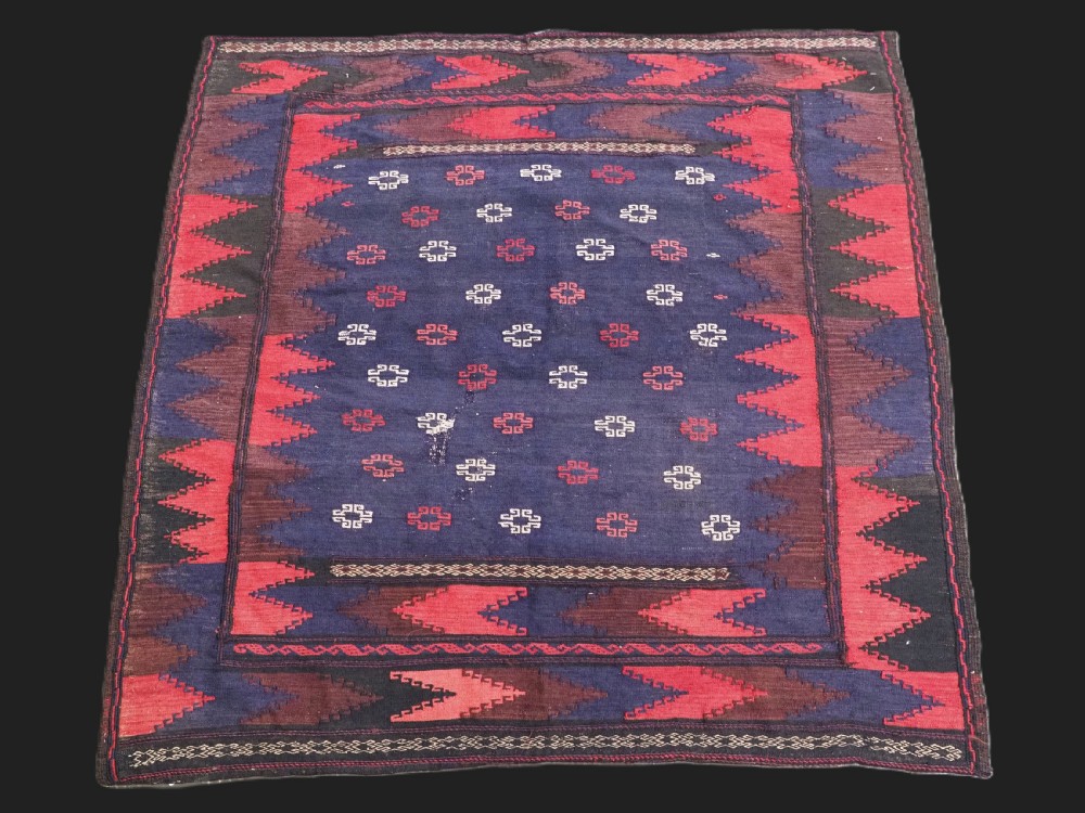 A rectangular antique Baluch sofreh flatweave textile featuring an indigo blue centre with small geometric motifs framed by serrated red and dark blue borders.