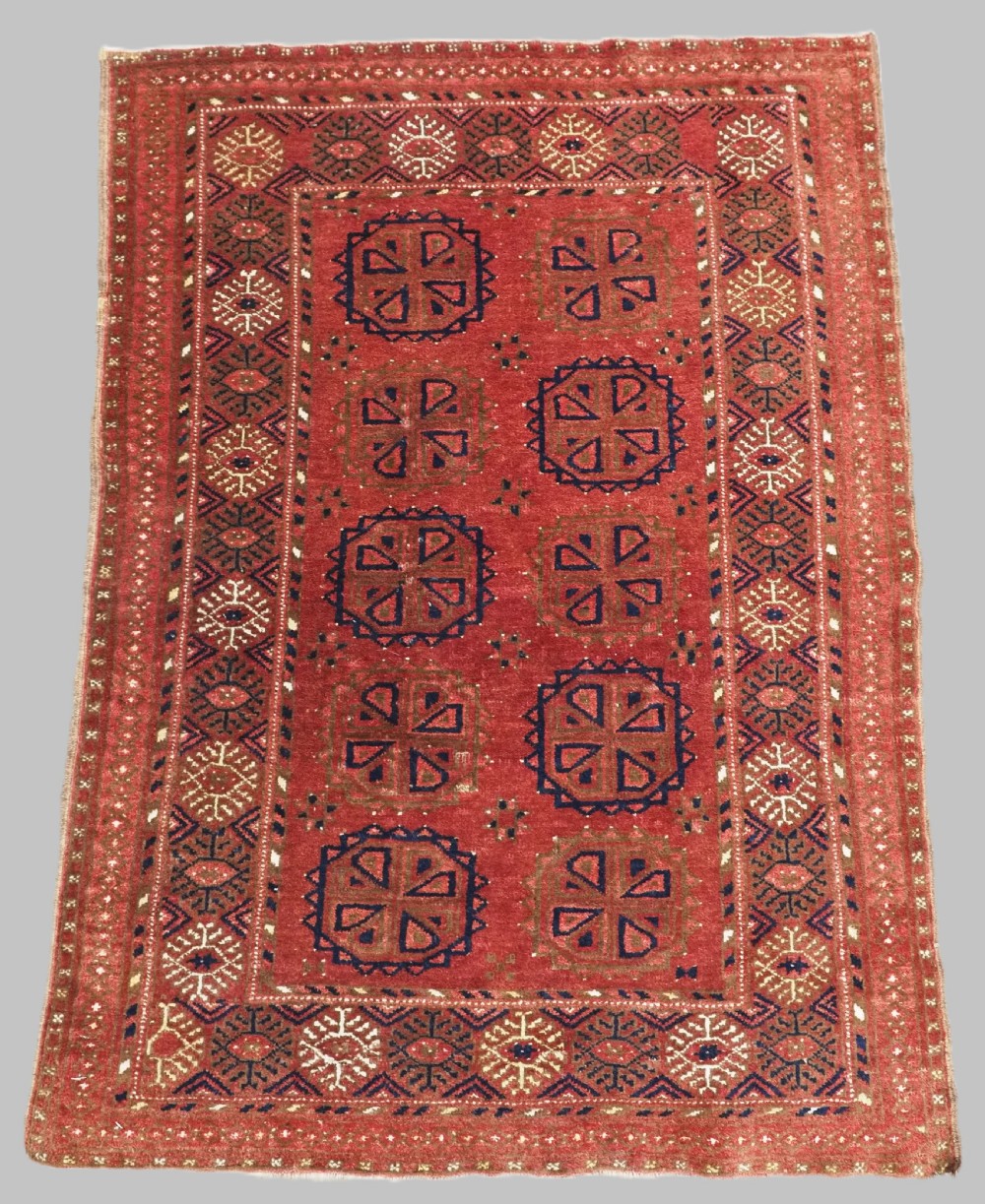 A rectangular hand-knotted wool rug featuring repeating octagonal gul motifs in dark blue and brown set against a deep red field with geometric patterned borders.
