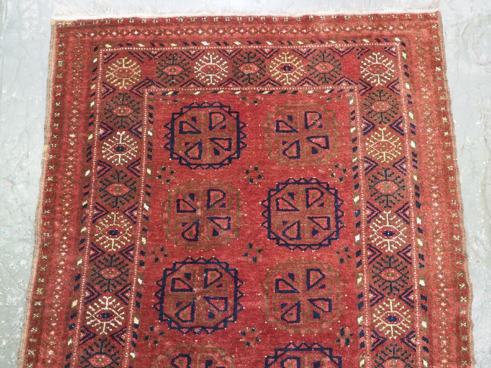 A rectangular hand-knotted wool rug featuring repeating octagonal gul motifs in dark blue and brown set against a deep red field with geometric patterned borders. View III
