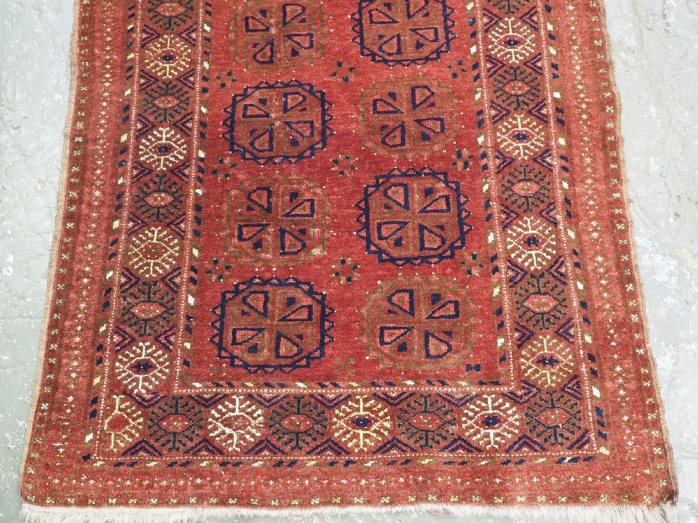 A rectangular hand-knotted wool rug featuring repeating octagonal gul motifs in dark blue and brown set against a deep red field with geometric patterned borders. View IV