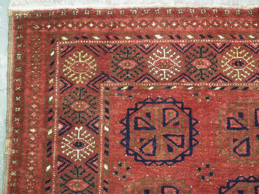 A rectangular hand-knotted wool rug featuring repeating octagonal gul motifs in dark blue and brown set against a deep red field with geometric patterned borders. View V