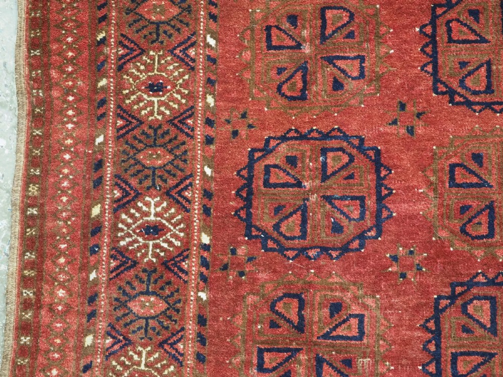 A rectangular hand-knotted wool rug featuring repeating octagonal gul motifs in dark blue and brown set against a deep red field with geometric patterned borders. View VI