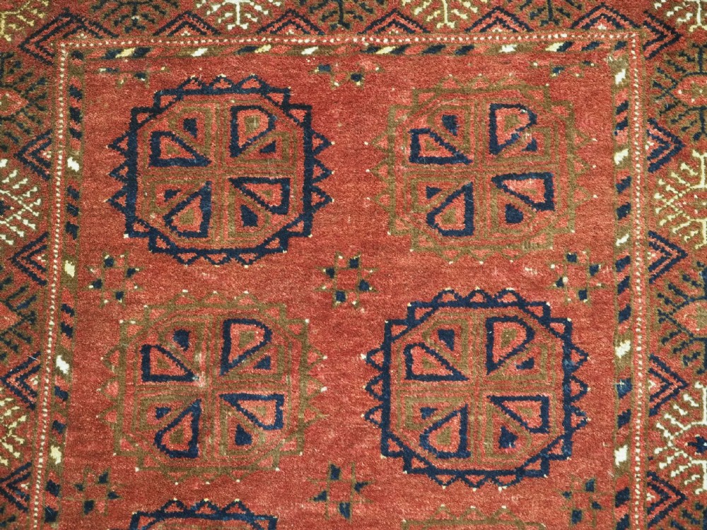 A rectangular hand-knotted wool rug featuring repeating octagonal gul motifs in dark blue and brown set against a deep red field with geometric patterned borders. View VII
