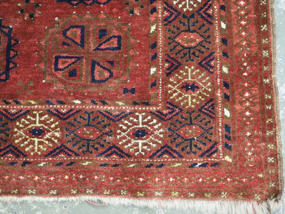 A rectangular hand-knotted wool rug featuring repeating octagonal gul motifs in dark blue and brown set against a deep red field with geometric patterned borders. View IX