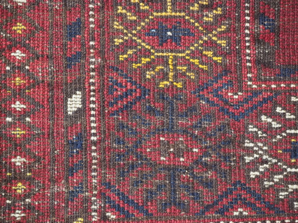 A rectangular hand-knotted wool rug featuring repeating octagonal gul motifs in dark blue and brown set against a deep red field with geometric patterned borders. View X