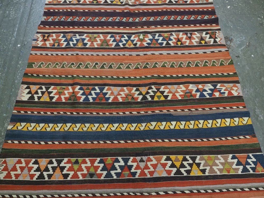 An antique handwoven wool Caucasian Shirvan kilim rug featuring horizontal bands filled with geometric serrated diamond and zig-zag patterns in terracotta, blue, green, and yellow tones. View IV