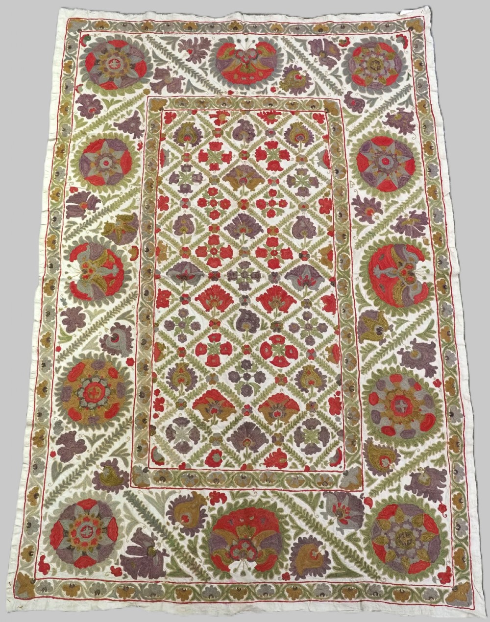 A rectangular embroidered suzani textile featuring a central diamond lattice pattern surrounded by a border of large floral medallions on a cream background.