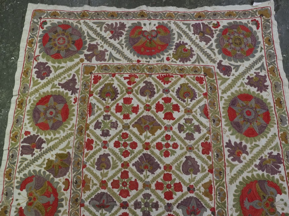 A rectangular embroidered suzani textile featuring a central diamond lattice pattern surrounded by a border of large floral medallions on a cream background. View III