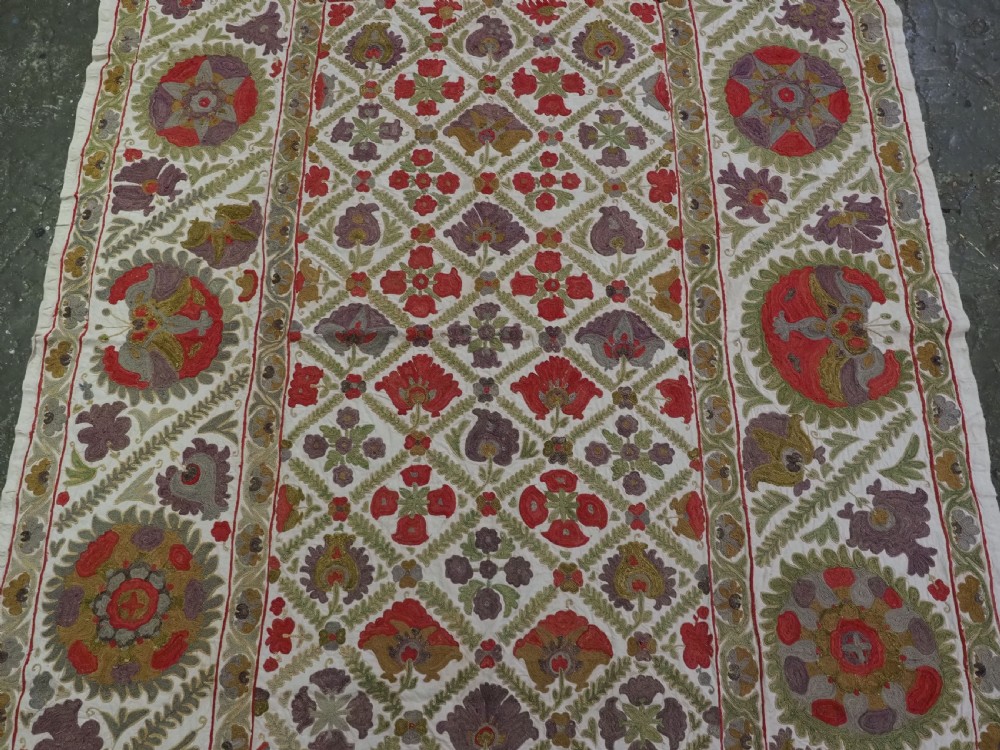 A rectangular embroidered suzani textile featuring a central diamond lattice pattern surrounded by a border of large floral medallions on a cream background. View IV