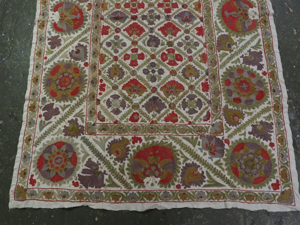 A rectangular embroidered suzani textile featuring a central diamond lattice pattern surrounded by a border of large floral medallions on a cream background. View V