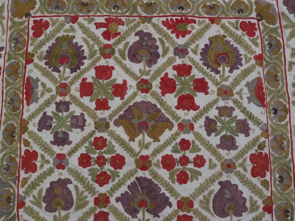 A rectangular embroidered suzani textile featuring a central diamond lattice pattern surrounded by a border of large floral medallions on a cream background. View IX