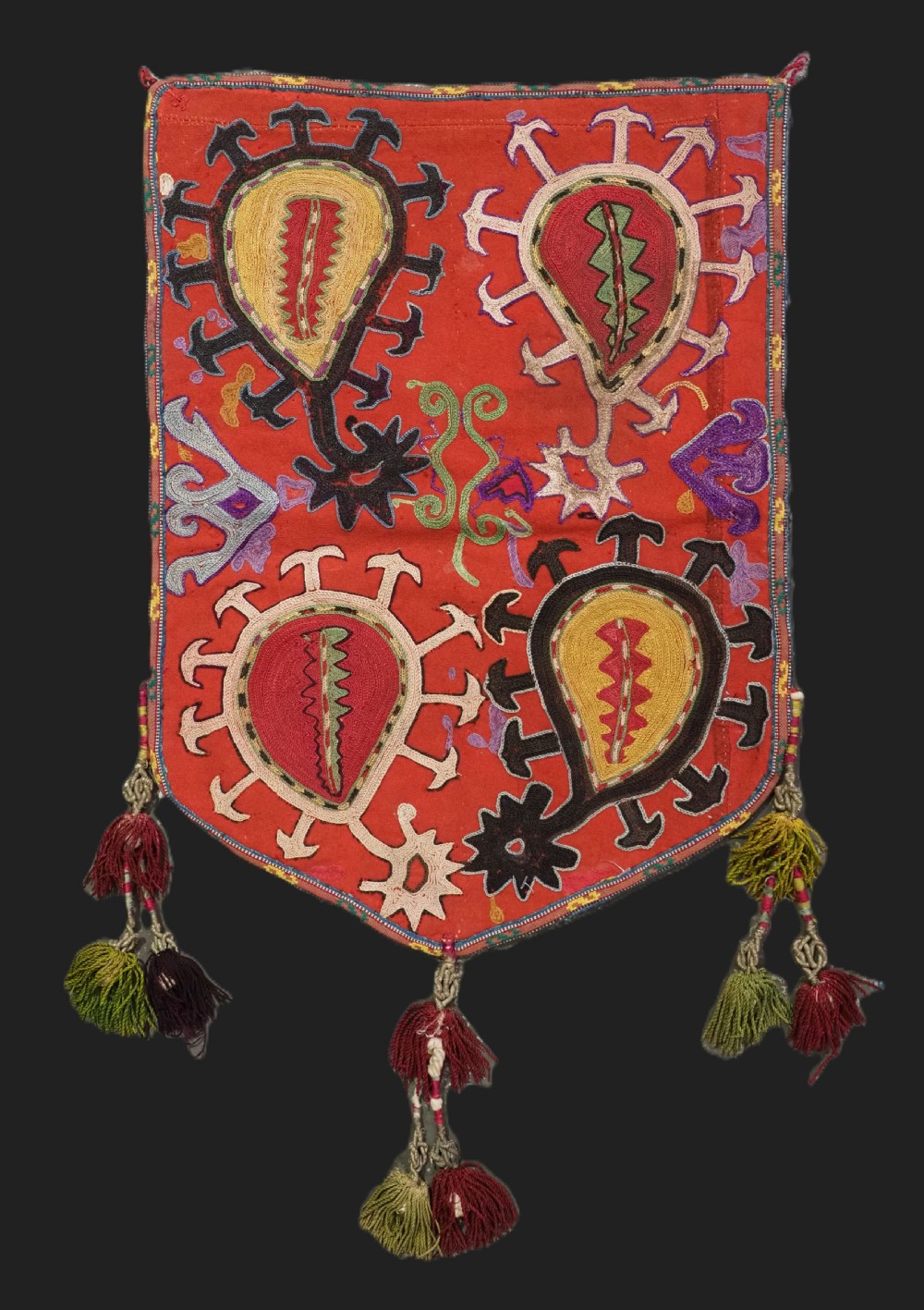 An antique Uzbek shield-shaped red wool textile embroidered with colourful silk teardrop motifs and finished with hanging wool tassels.