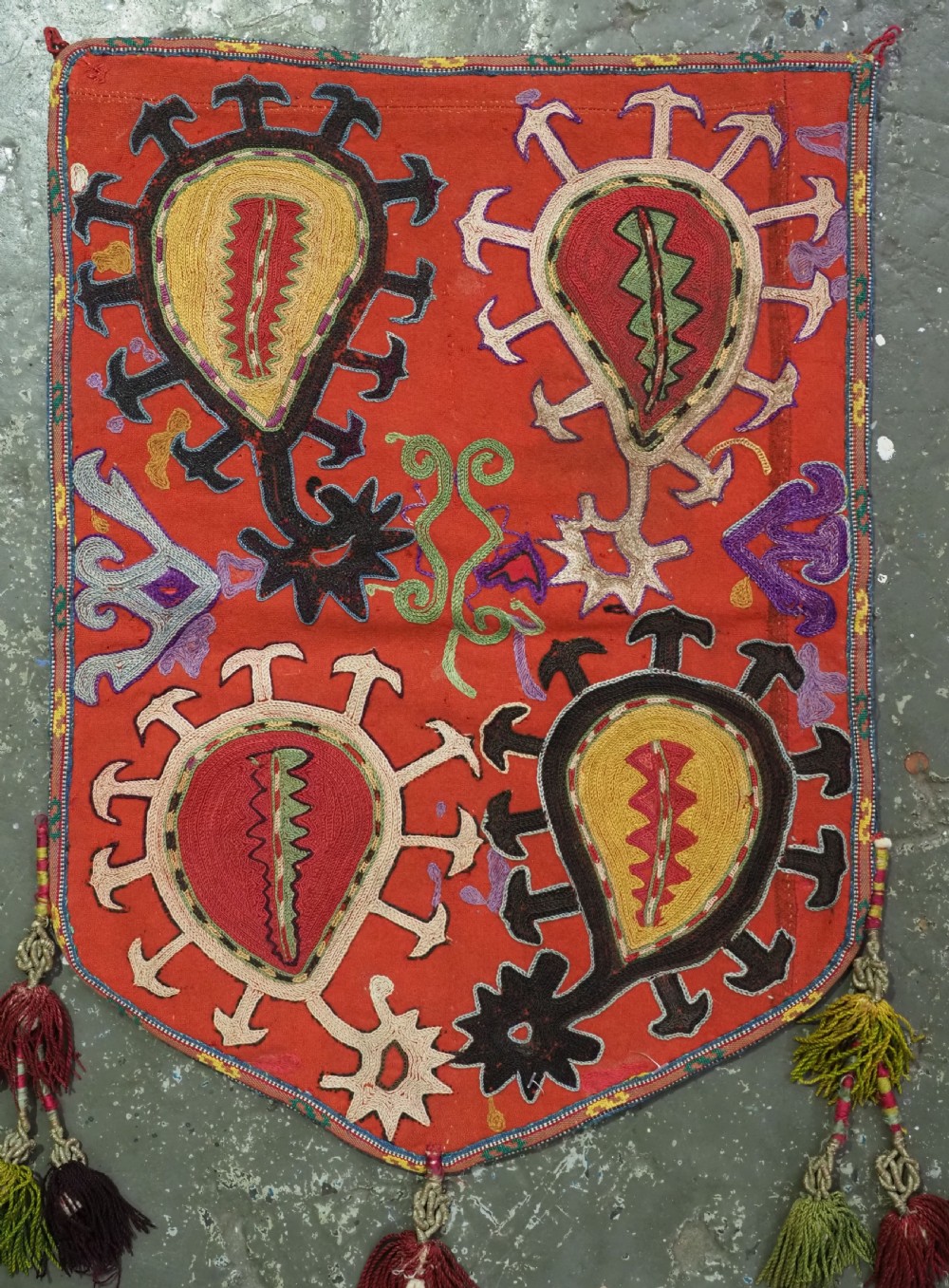 An antique Uzbek shield-shaped red wool textile embroidered with colourful silk teardrop motifs and finished with hanging wool tassels. View X