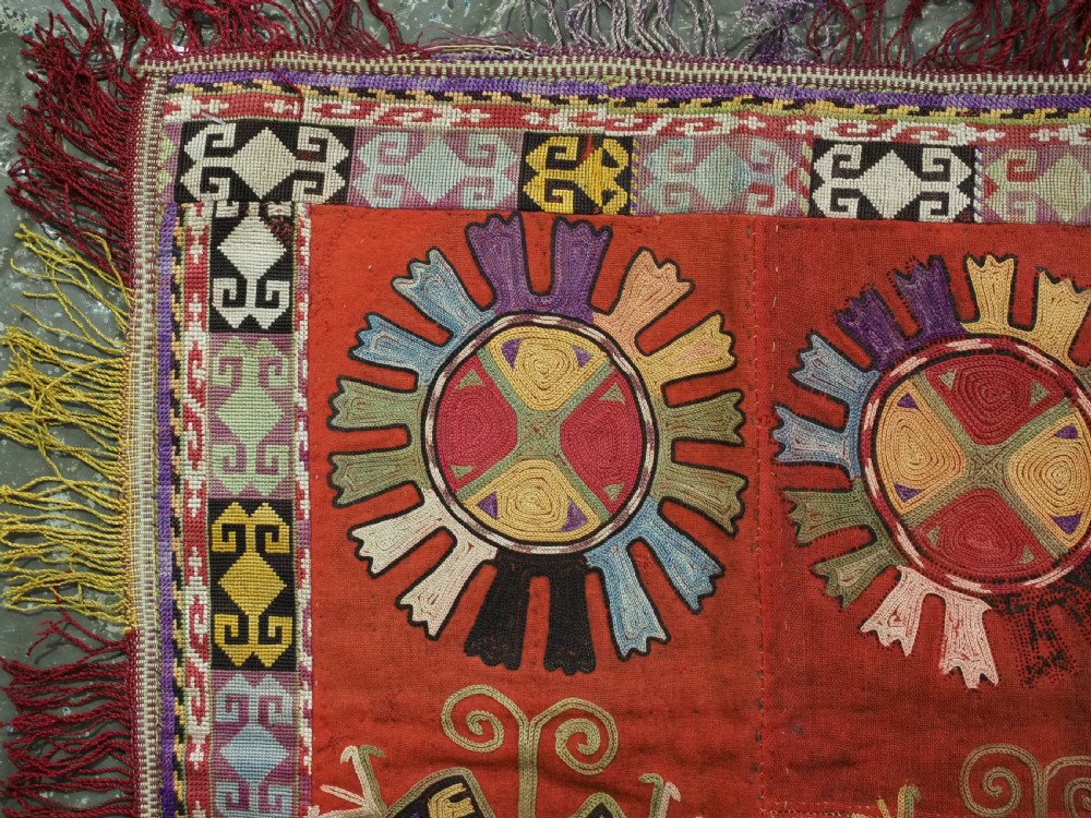 A square red embroidered Uzbek Lakai tribal ilgich wall hanging featuring solar medallions, geometric borders, and coloured fringe. View II