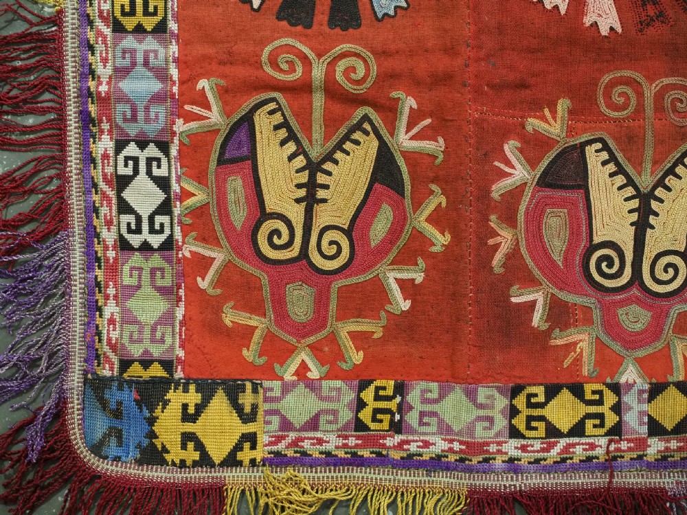 A square red embroidered Uzbek Lakai tribal ilgich wall hanging featuring solar medallions, geometric borders, and coloured fringe. View III