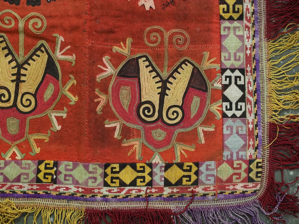 A square red embroidered Uzbek Lakai tribal ilgich wall hanging featuring solar medallions, geometric borders, and coloured fringe. View IV