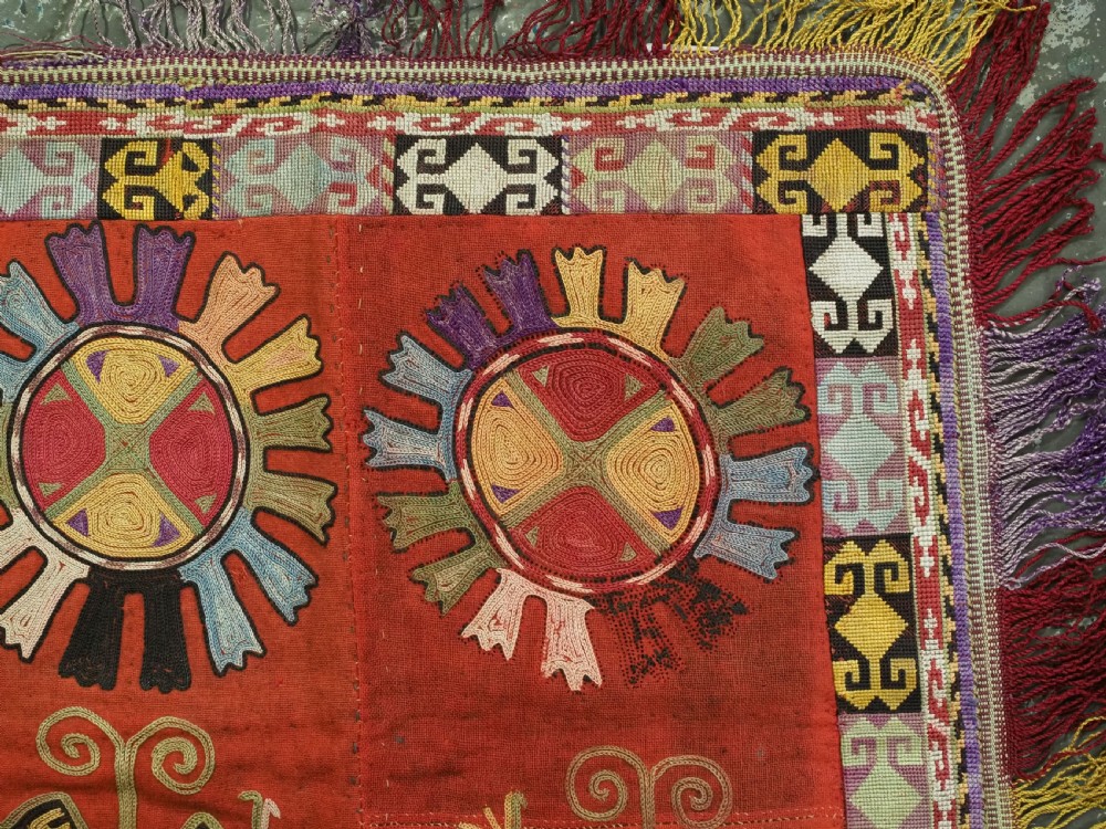 A square red embroidered Uzbek Lakai tribal ilgich wall hanging featuring solar medallions, geometric borders, and coloured fringe. View V
