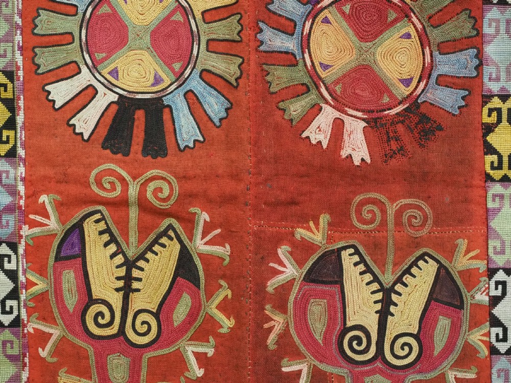 A square red embroidered Uzbek Lakai tribal ilgich wall hanging featuring solar medallions, geometric borders, and coloured fringe. View VI