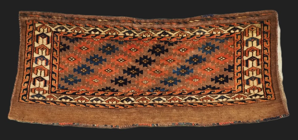 An antique woven Yomut Turkmen torba displaying repeated geometric Aishk gul patterns in red, blue, and ivory on a warm brown background.