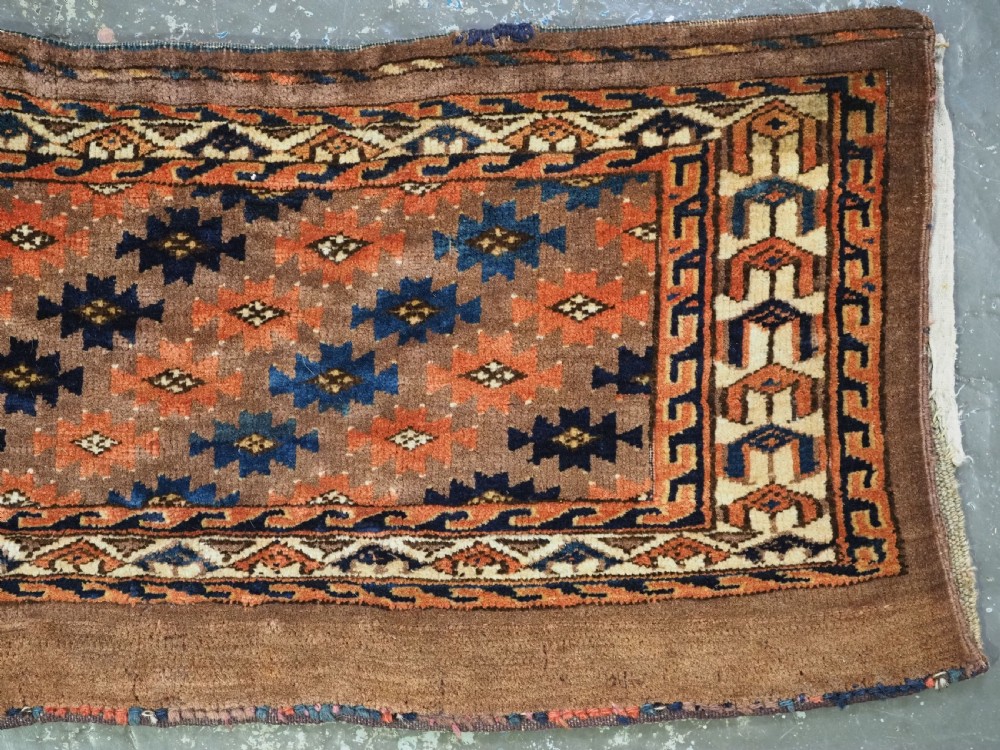 An antique woven Yomut Turkmen torba displaying repeated geometric Aishk gul patterns in red, blue, and ivory on a warm brown background. View IV