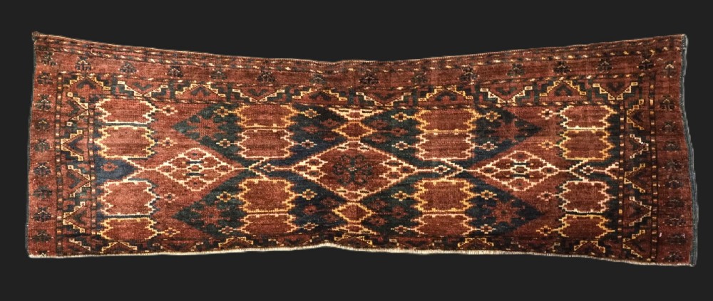 A horizontal hand-knotted antique wool torba displaying a repeating ikat diamond pattern in terracotta red, navy blue, and ochre hues.