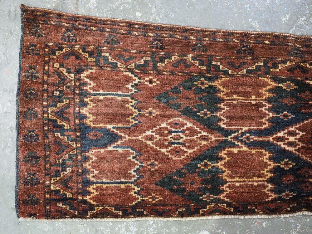 A horizontal hand-knotted antique wool torba displaying a repeating ikat diamond pattern in terracotta red, navy blue, and ochre hues. View II