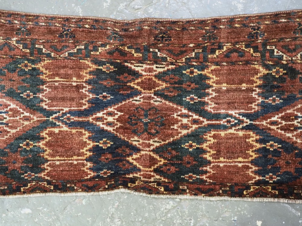 A horizontal hand-knotted antique wool torba displaying a repeating ikat diamond pattern in terracotta red, navy blue, and ochre hues. View III