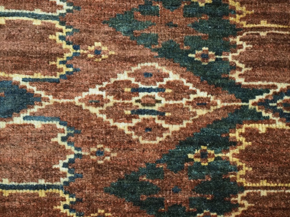 A horizontal hand-knotted antique wool torba displaying a repeating ikat diamond pattern in terracotta red, navy blue, and ochre hues. View V