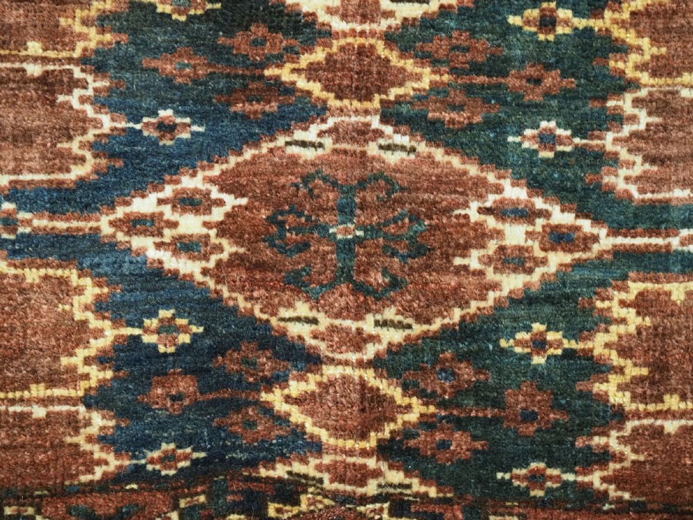 A horizontal hand-knotted antique wool torba displaying a repeating ikat diamond pattern in terracotta red, navy blue, and ochre hues. View VI