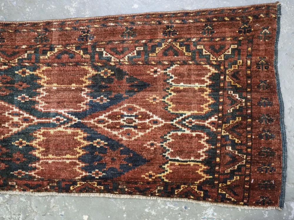 A horizontal hand-knotted antique wool torba displaying a repeating ikat diamond pattern in terracotta red, navy blue, and ochre hues. View X