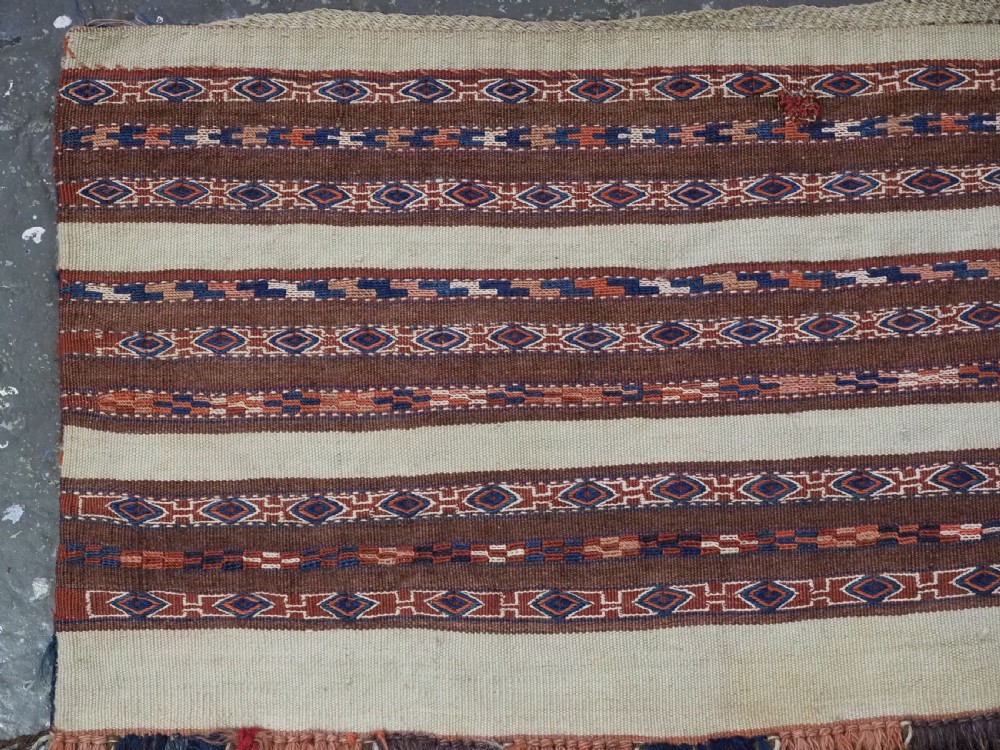 An antique hand-woven Turkmen flatweave textile panel featuring horizontal bands of geometric motifs in terracotta, dark blue, and brown on a cream background with long fringed trim. View II