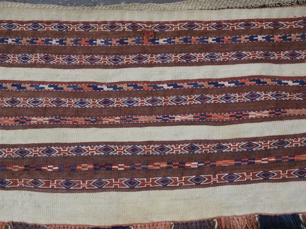 An antique hand-woven Turkmen flatweave textile panel featuring horizontal bands of geometric motifs in terracotta, dark blue, and brown on a cream background with long fringed trim. View III