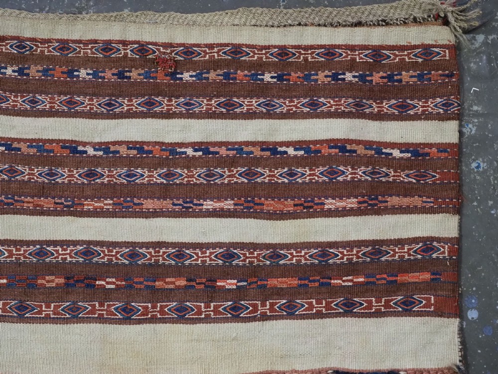 An antique hand-woven Turkmen flatweave textile panel featuring horizontal bands of geometric motifs in terracotta, dark blue, and brown on a cream background with long fringed trim. View IV