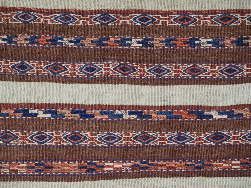 An antique hand-woven Turkmen flatweave textile panel featuring horizontal bands of geometric motifs in terracotta, dark blue, and brown on a cream background with long fringed trim. View V