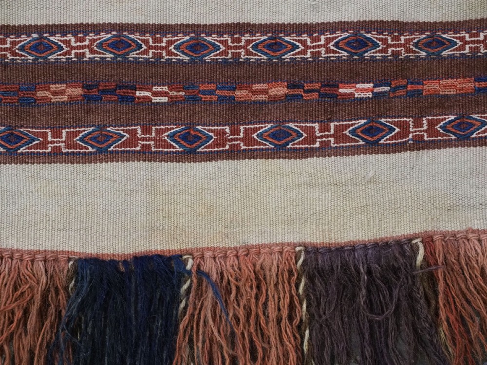 An antique hand-woven Turkmen flatweave textile panel featuring horizontal bands of geometric motifs in terracotta, dark blue, and brown on a cream background with long fringed trim. View VI