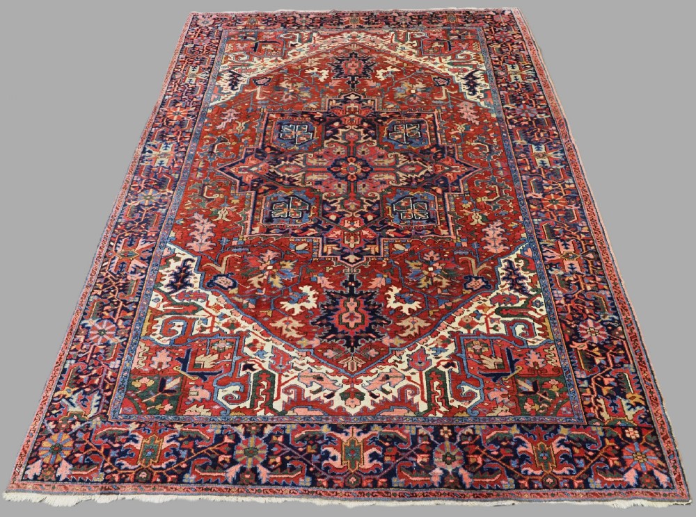 An antique hand-knotted Persian Heriz wool carpet featuring a central geometric medallion, angular spandrels, a terracotta red field, and navy blue borders.