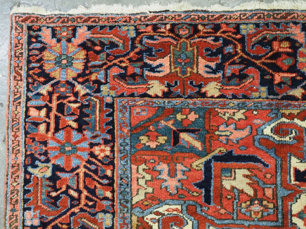 An antique hand-knotted Persian Heriz wool carpet featuring a central geometric medallion, angular spandrels, a terracotta red field, and navy blue borders. View V