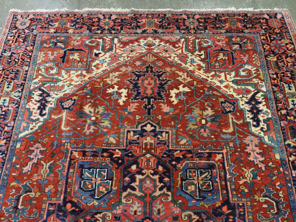 An antique hand-knotted Persian Heriz wool carpet featuring a central geometric medallion, angular spandrels, a terracotta red field, and navy blue borders. View VI