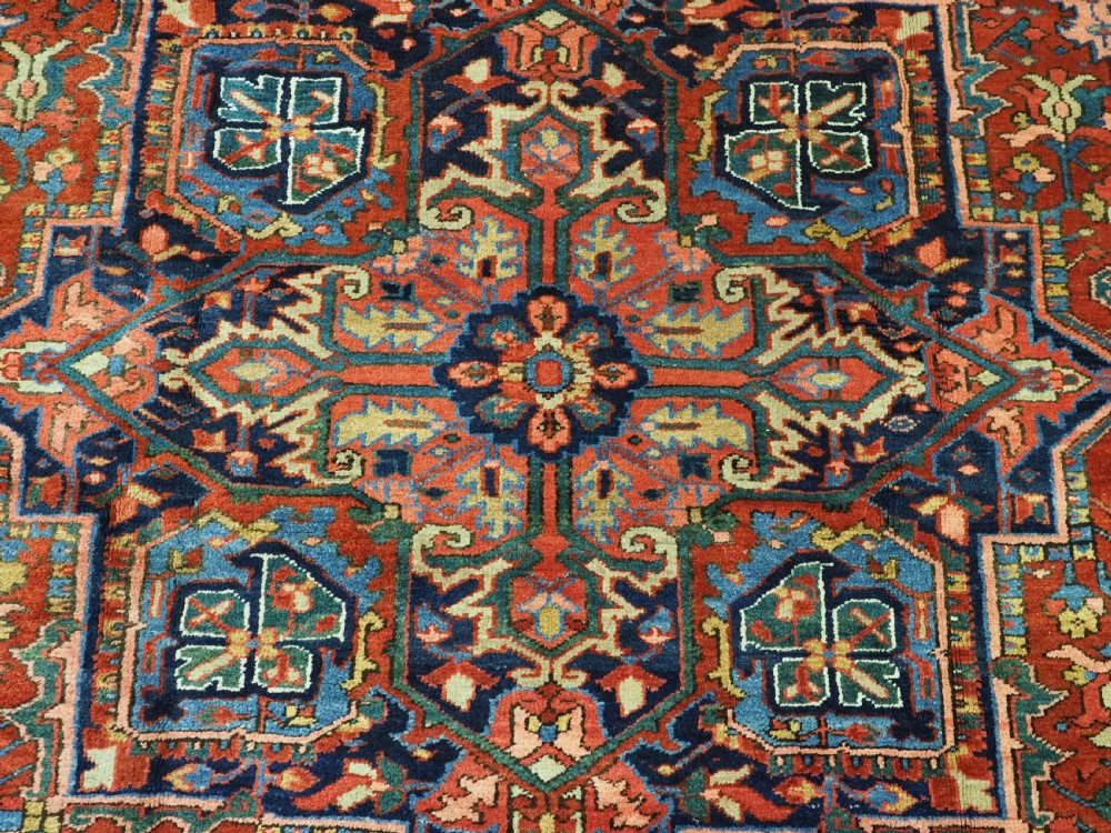 An antique hand-knotted Persian Heriz wool carpet featuring a central geometric medallion, angular spandrels, a terracotta red field, and navy blue borders. View IX