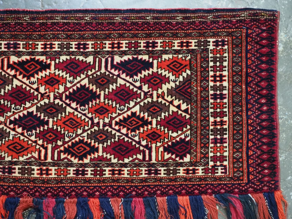 An antique rectangular Tekke Turkmen woven torba face featuring a geometric diamond lattice pattern in burgundy, cream, and orange, finished with long polychrome wool fringes along the lower edge. View V