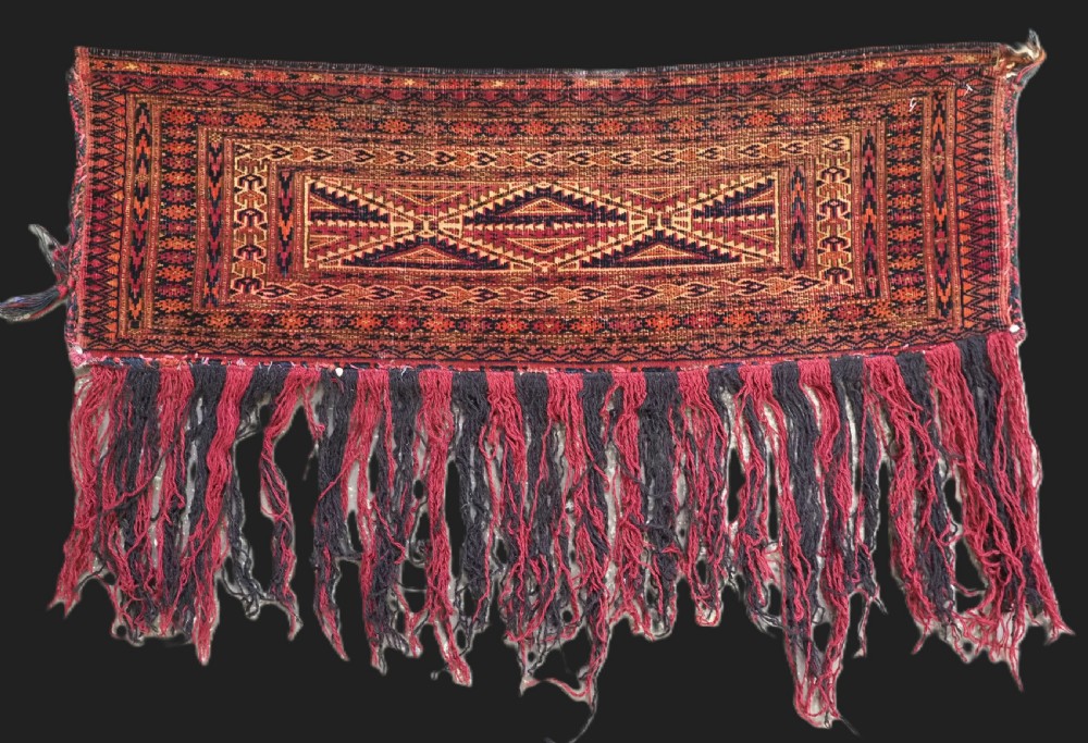 An antique woven Tekke Turkmen torba featuring intricate geometric lattice motifs in dark red, terracotta, and dark blue with a long braided fringe along the bottom edge.