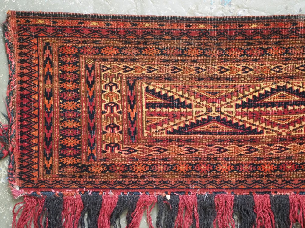 An antique woven Tekke Turkmen torba featuring intricate geometric lattice motifs in dark red, terracotta, and dark blue with a long braided fringe along the bottom edge. View III