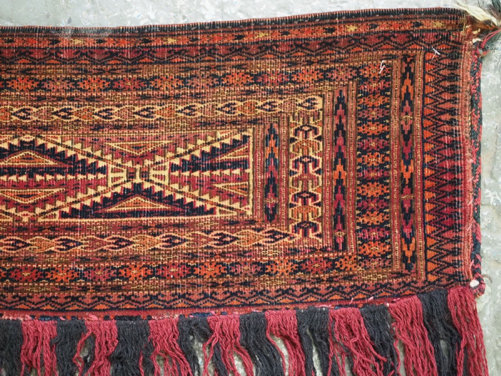 An antique woven Tekke Turkmen torba featuring intricate geometric lattice motifs in dark red, terracotta, and dark blue with a long braided fringe along the bottom edge. View IV