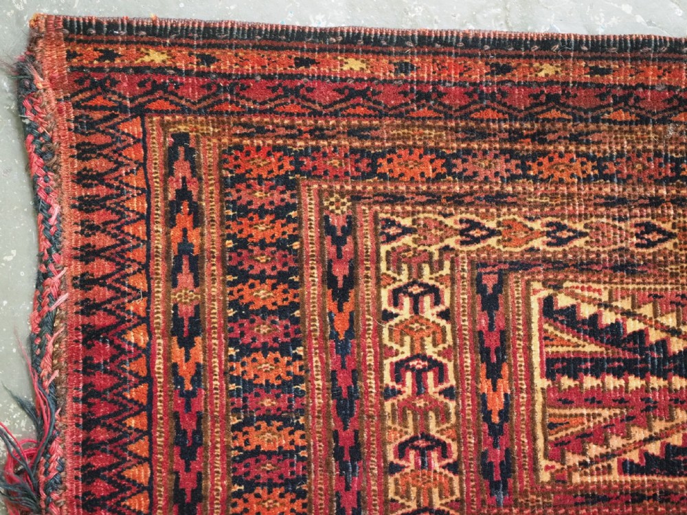 An antique woven Tekke Turkmen torba featuring intricate geometric lattice motifs in dark red, terracotta, and dark blue with a long braided fringe along the bottom edge. View V