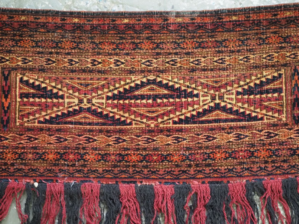 An antique woven Tekke Turkmen torba featuring intricate geometric lattice motifs in dark red, terracotta, and dark blue with a long braided fringe along the bottom edge. View VI