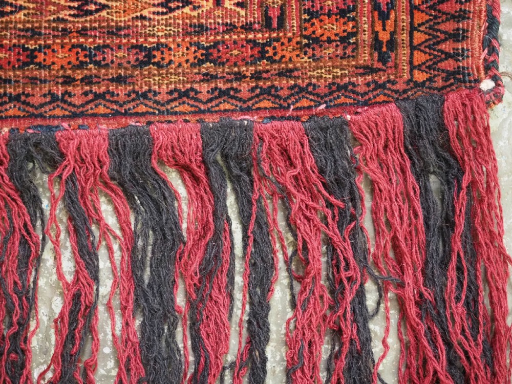 An antique woven Tekke Turkmen torba featuring intricate geometric lattice motifs in dark red, terracotta, and dark blue with a long braided fringe along the bottom edge. View IX